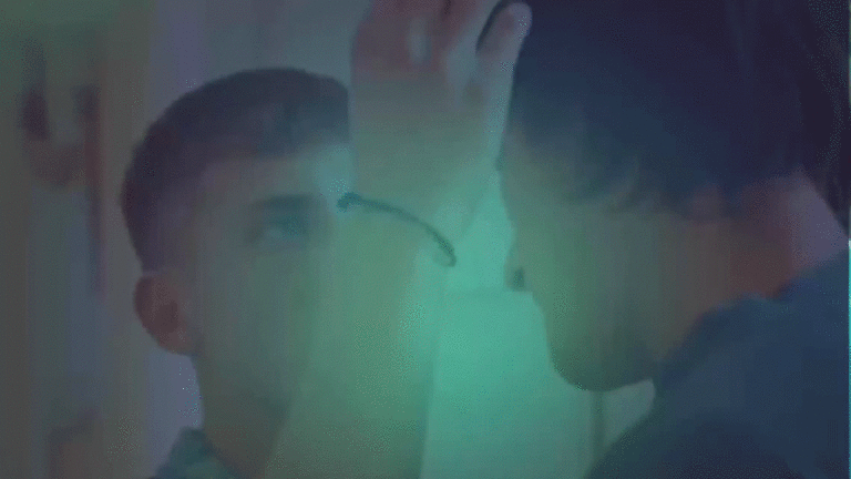 Two individuals in a close interaction, with one person touching the other's forehead in a dimly lit setting.