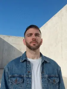Man wearing a denim jacket and white shirt standing in front of a concrete wall under a clear blue sky.