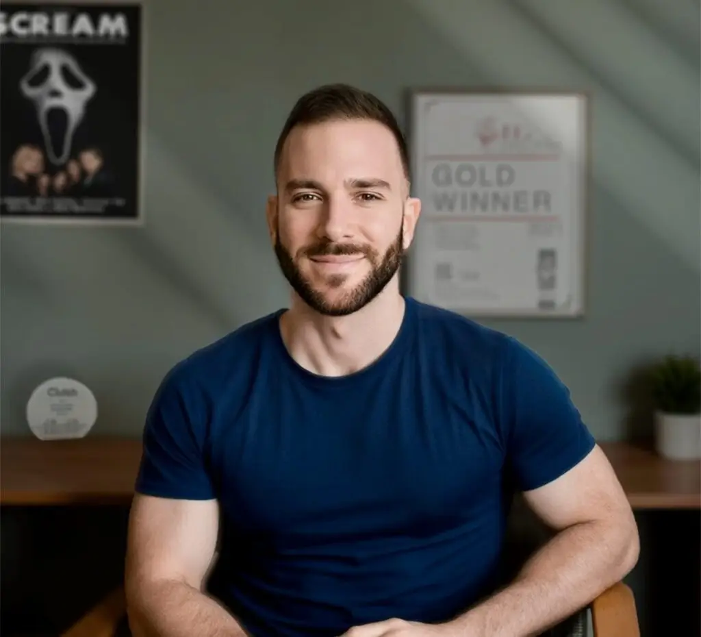 Man with a beard wearing a blue t-shirt, seated in an office chair, smiling at the camera. Man with a beard wearing a blue t-shirt, seated in an office chair, smiling at the camera.