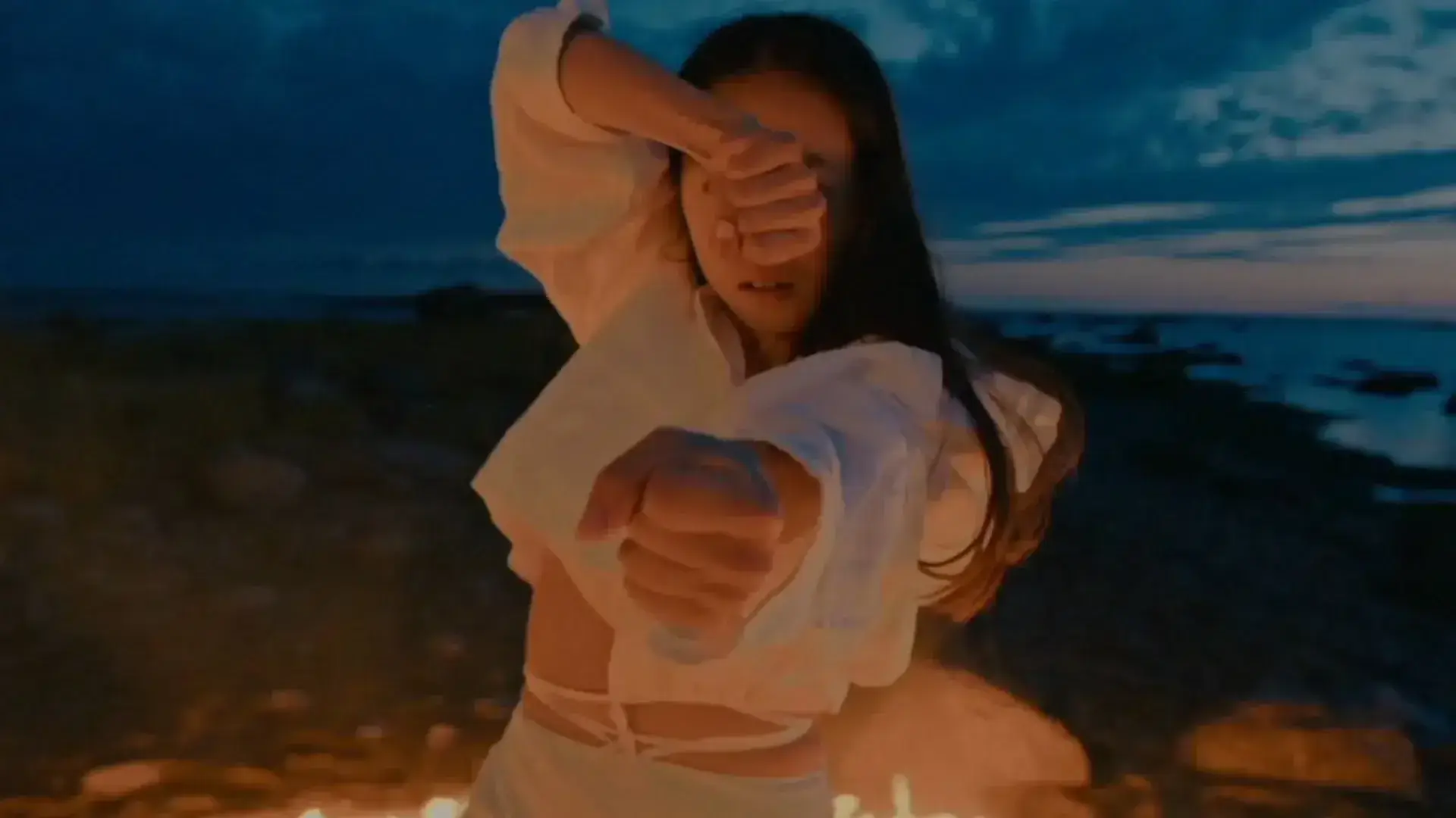 A woman in a white outfit performing a dance with fire at dusk, with a dramatic sky in the background.