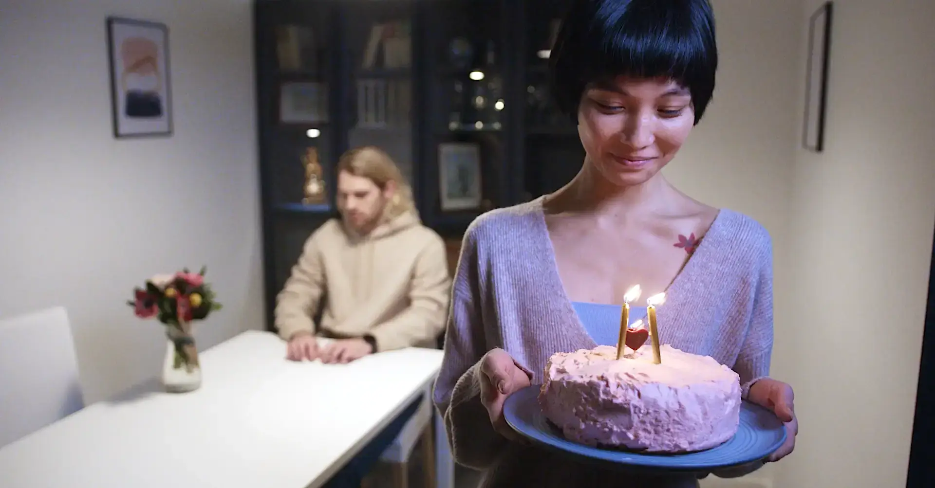 Woman with short hair holding a pink birthday cake with two lit candles while a man sits at a table in the background.
