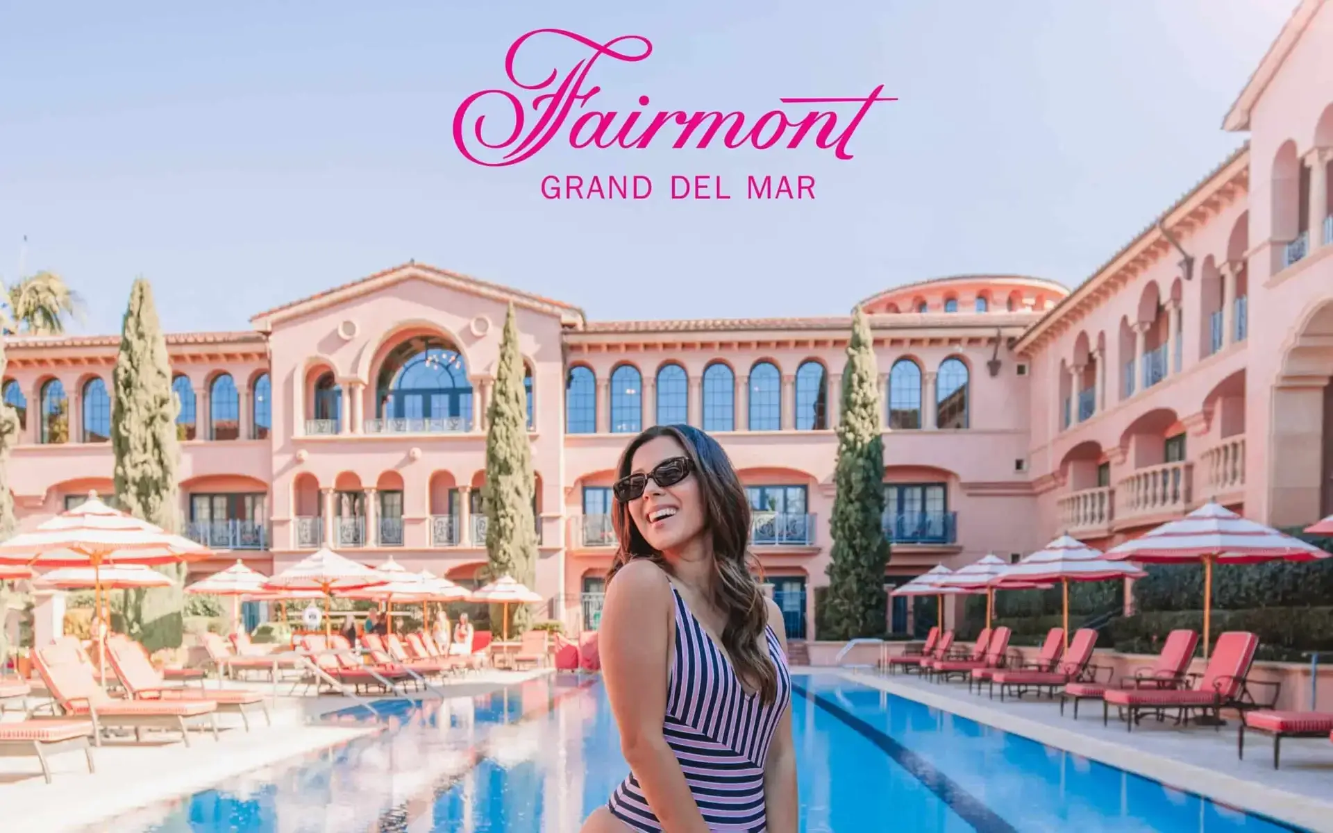 Woman in sunglasses posing by the pool at Fairmont Grand Del Mar resort with pink building in the background.