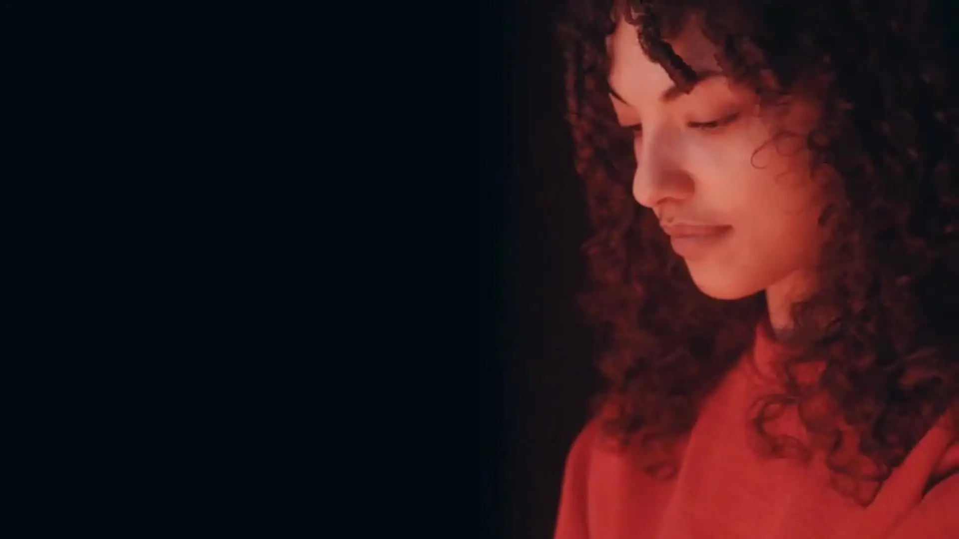 Profile view of a woman with curly hair wearing a red sweater, looking down with a neutral expression.