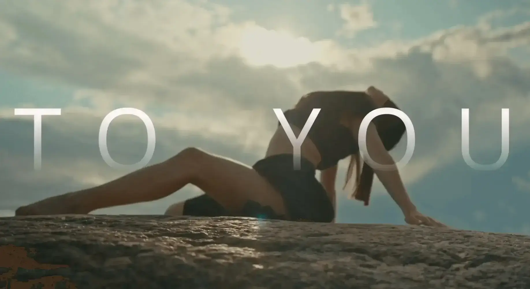 Woman performing an athletic pose on a rock under a cloudy sky with sunlight in the background.