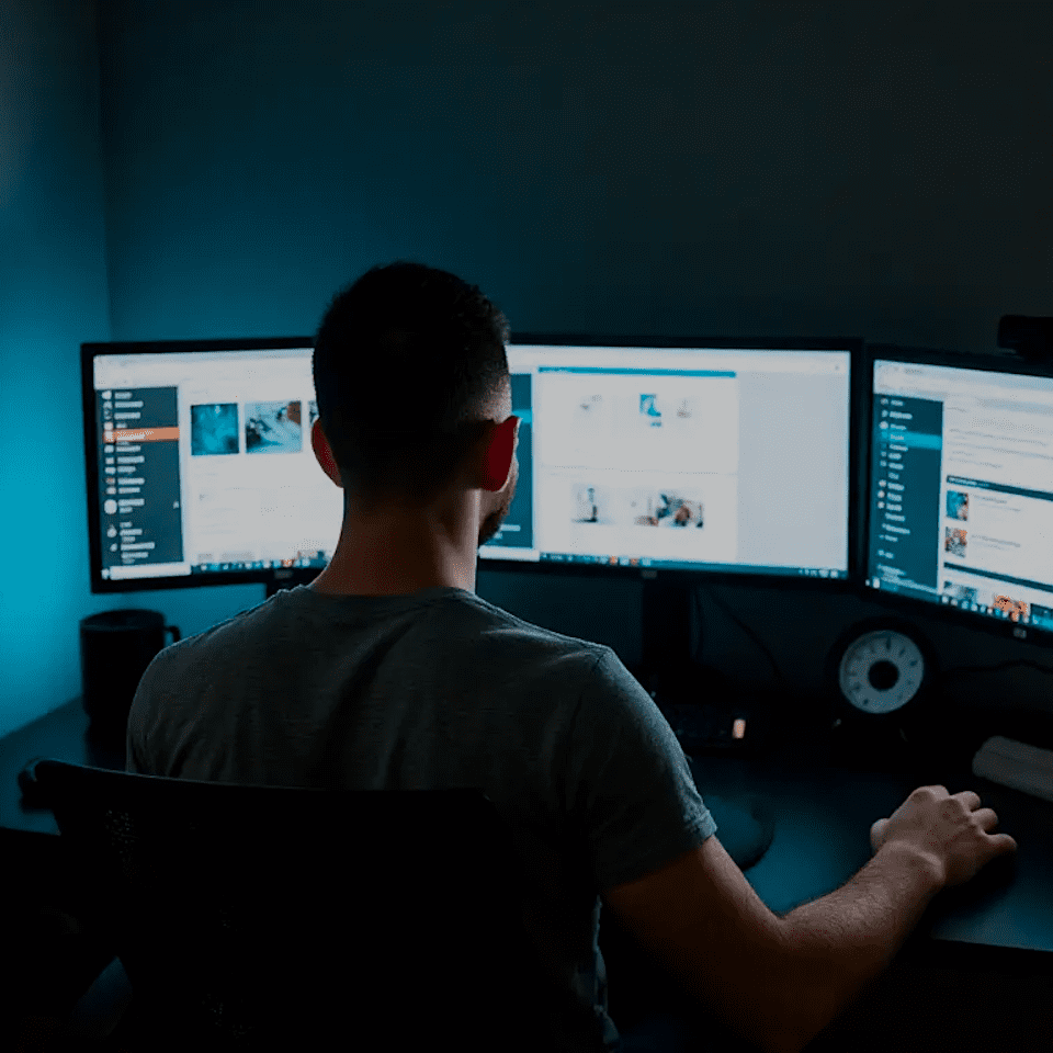 Individual seated at a desk using two computer monitors in a dimly lit room.